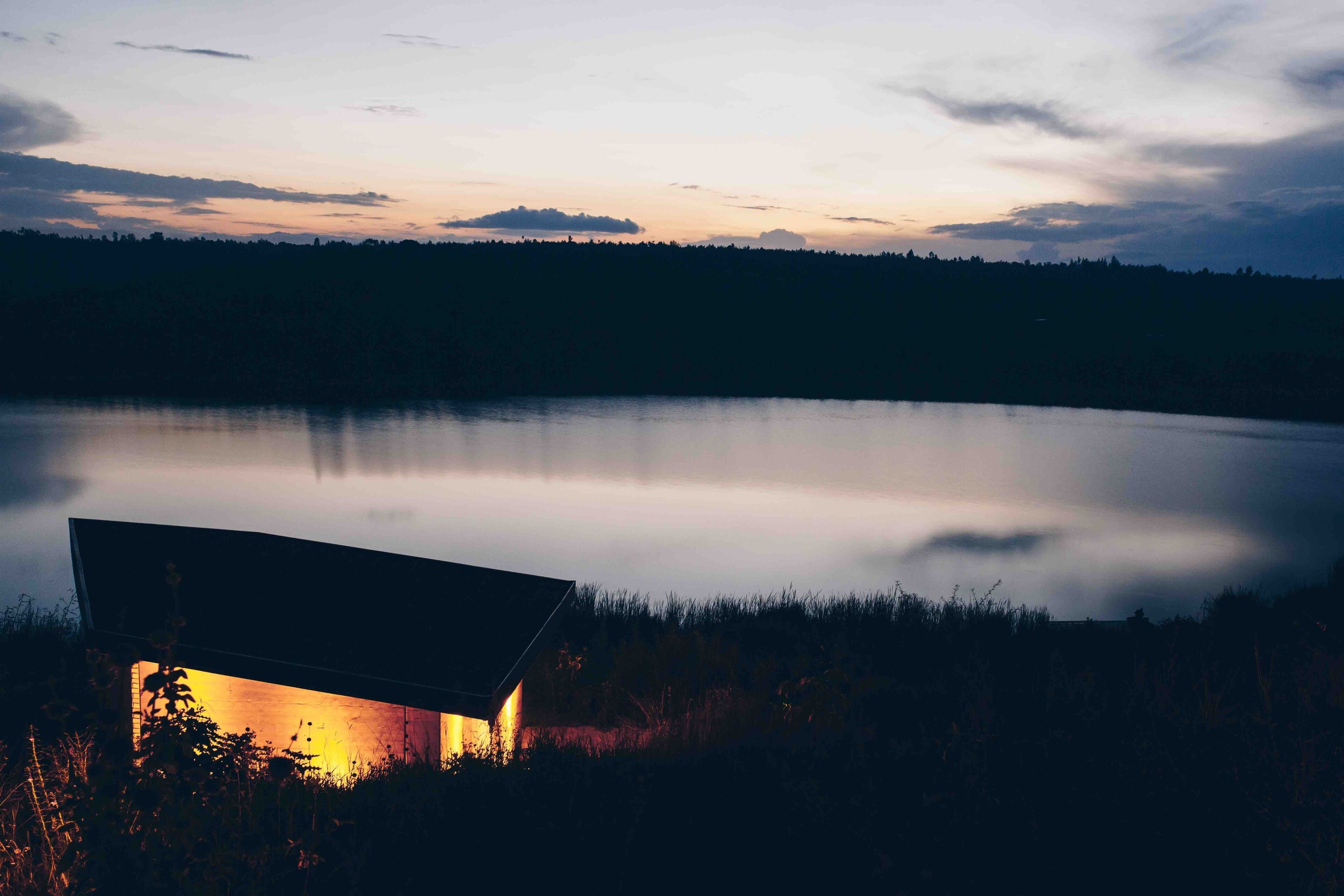 Una casa junto a un lago tranquilo al anochecer, con un techo visible e interior iluminado.