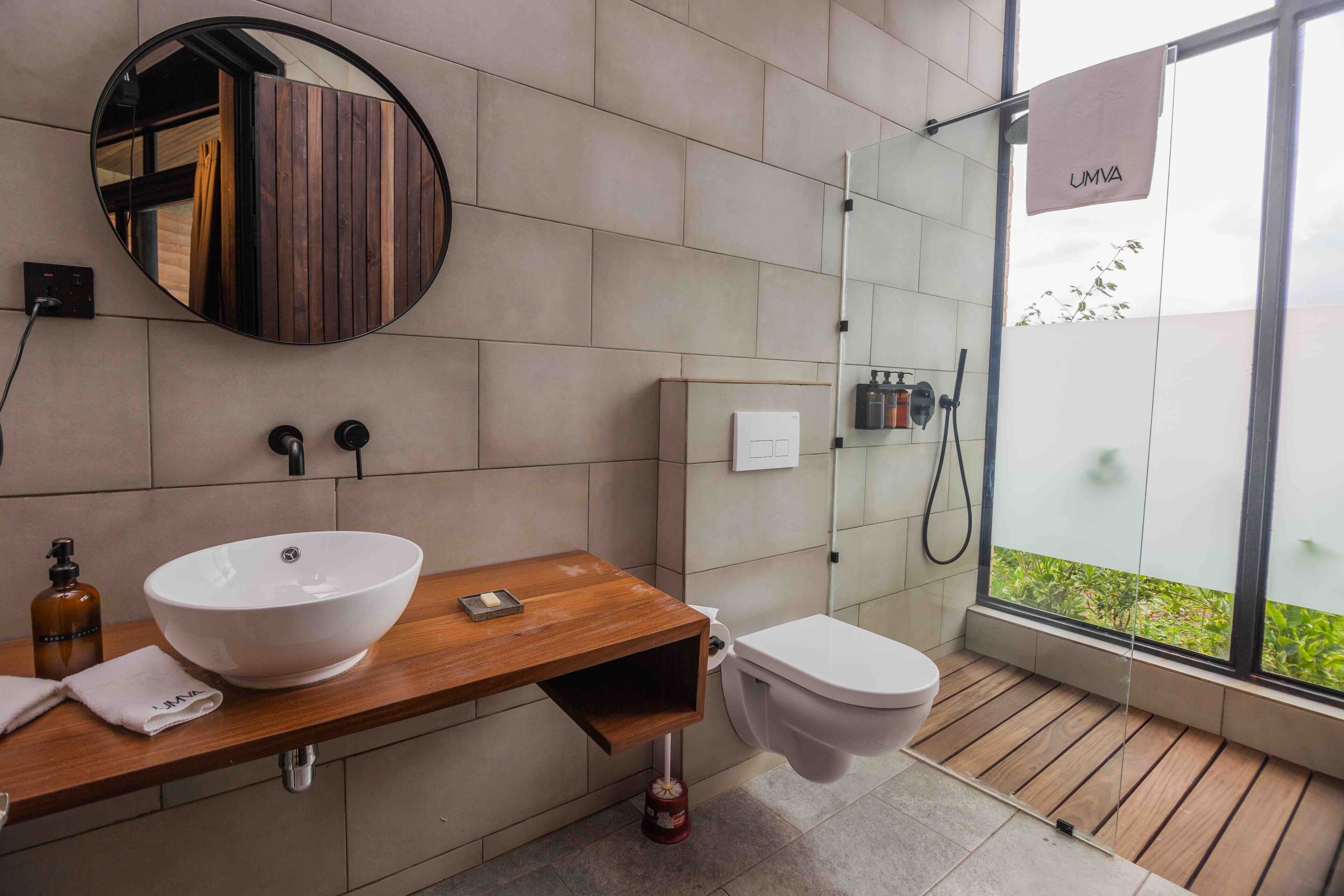 A modern bathroom with a wooden vanity, a large round mirror, a white sink, and a glass-enclosed shower.