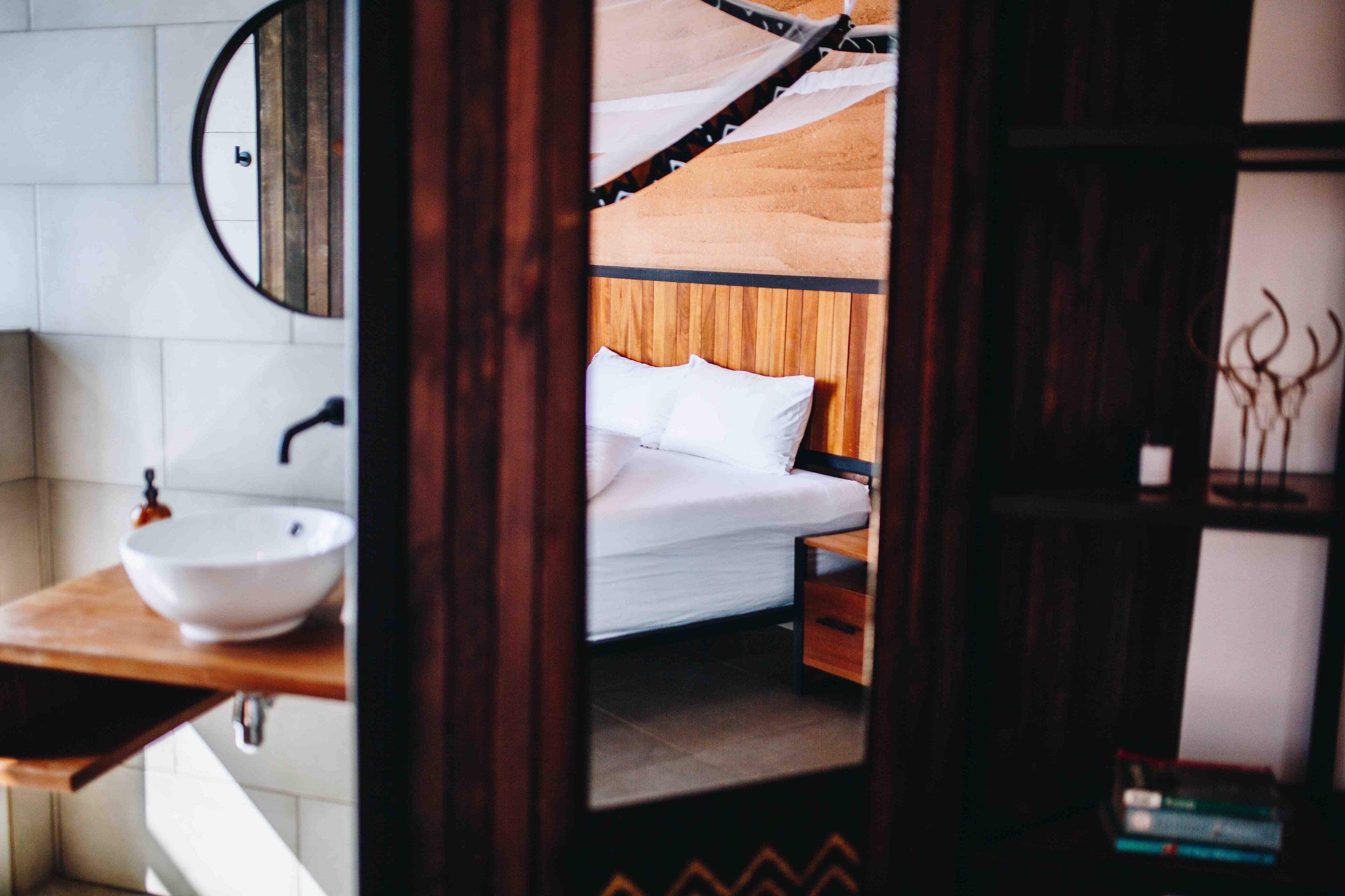 Un dormitorio con cama de madera, ropa de cama blanca, una mesita de noche y un espejo redondo en la pared.