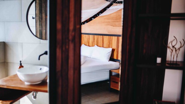 A bedroom with a wooden bed, white bedding, a bedside table, and a round mirror on the wall. - UMVA Muhazi (Muhazi)