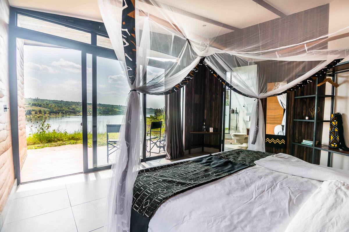 A modern bedroom with a large bed, sheer curtains, and a view of a lake.