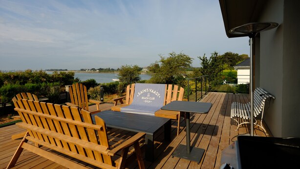 Eine Holzterrasse mit Adirondack-Stühlen und einem Tisch, mit Blick auf ein Gewässer und Grünflächen.