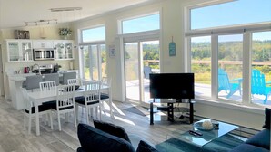Un vaste espace de vie comprenant une table à manger, des chaises, un canapé, un téléviseur et de grandes fenêtres donnant sur l’extérieur.