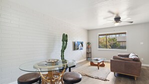 A modern living room with a glass dining table, two wicker chairs, a brown sofa, a ceiling fan, and a window with blinds. - Viridian Biltmore Villa with Pool (Phoenix)