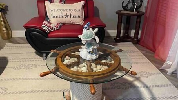 A glass-top table with a ship's wheel centerpiece, a red sofa with a 'Welcome to Our Beach House' pillow, and a small wooden side table with a decorative item. - Bacolet Beach Cottage-Luxe Pool (Palm Beach Gardens)