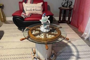 Een tafel met een glazen blad, versierd met een schipskoppel, een rode bank met een kussen waarop staat 'Welkom in Ons Strandhuis', en een klein houten bijzettafeltje met een decoratief object.