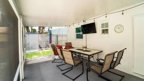 A covered patio with a table and chairs, a TV mounted on the wall, and a clock. - Bacolet Beach Cottage-Luxe Pool (Palm Beach Gardens)
