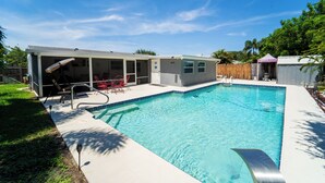 A rectangular swimming pool with a tiled edge, a covered patio area with a table and chairs, and a small shed in the background. - Bacolet Beach Cottage-Luxe Pool (Palm Beach Gardens)
