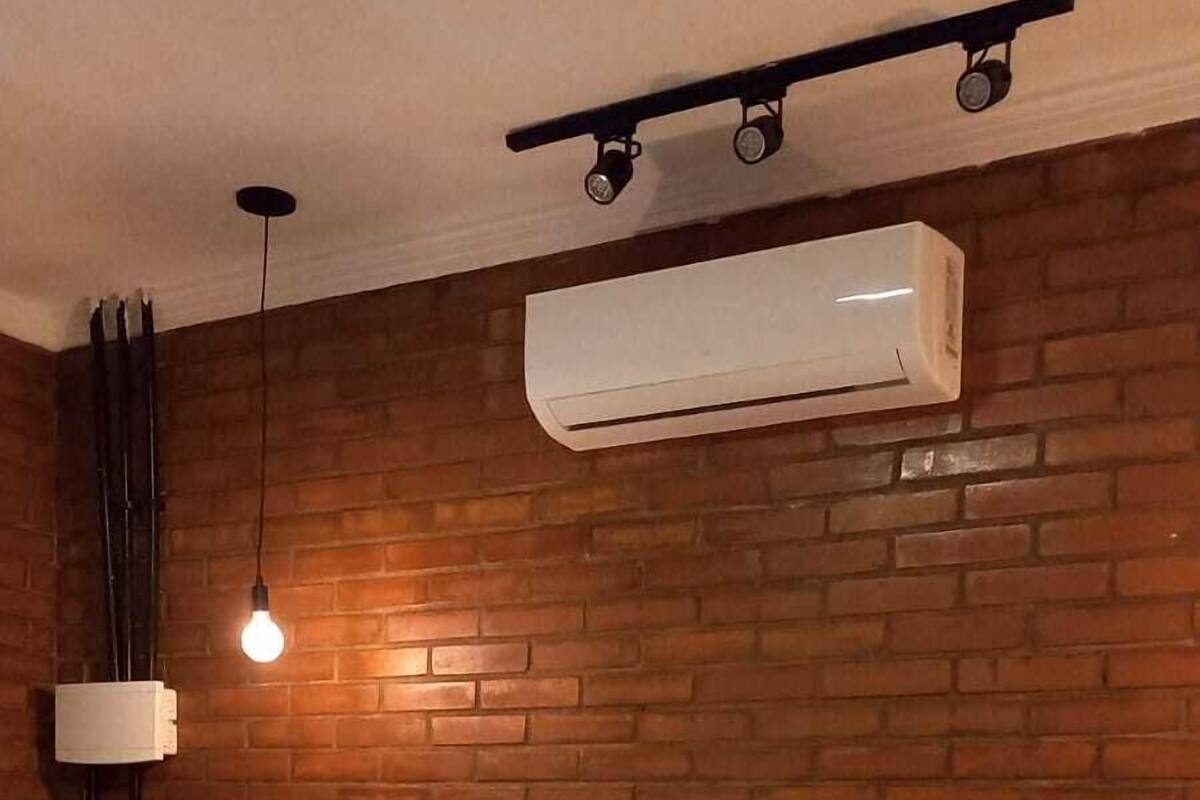 A bedroom with a brick wall, a bed with white bedding, a hanging light fixture, and an air conditioning unit.