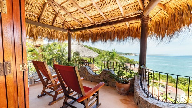 A wooden rocking chair and a stone wall on a balcony with a thatched roof, overlooking a coastal view. - Casa Ceiba Huatulco - Adults Only (Santa María Huatulco)
