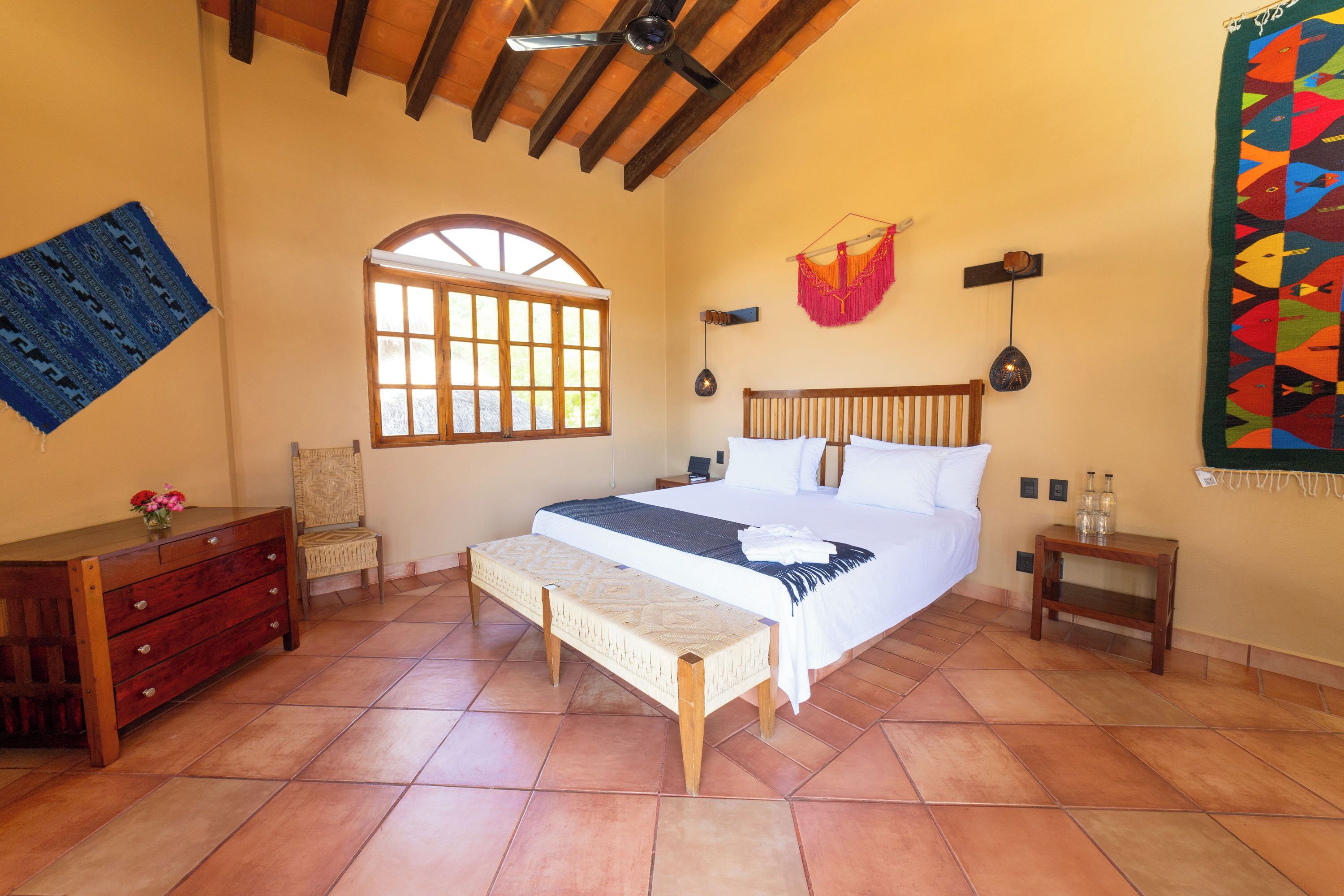 A bedroom with a bed, wooden headboard, bedside tables, a bench, a dresser, a window, and a wall hanging.