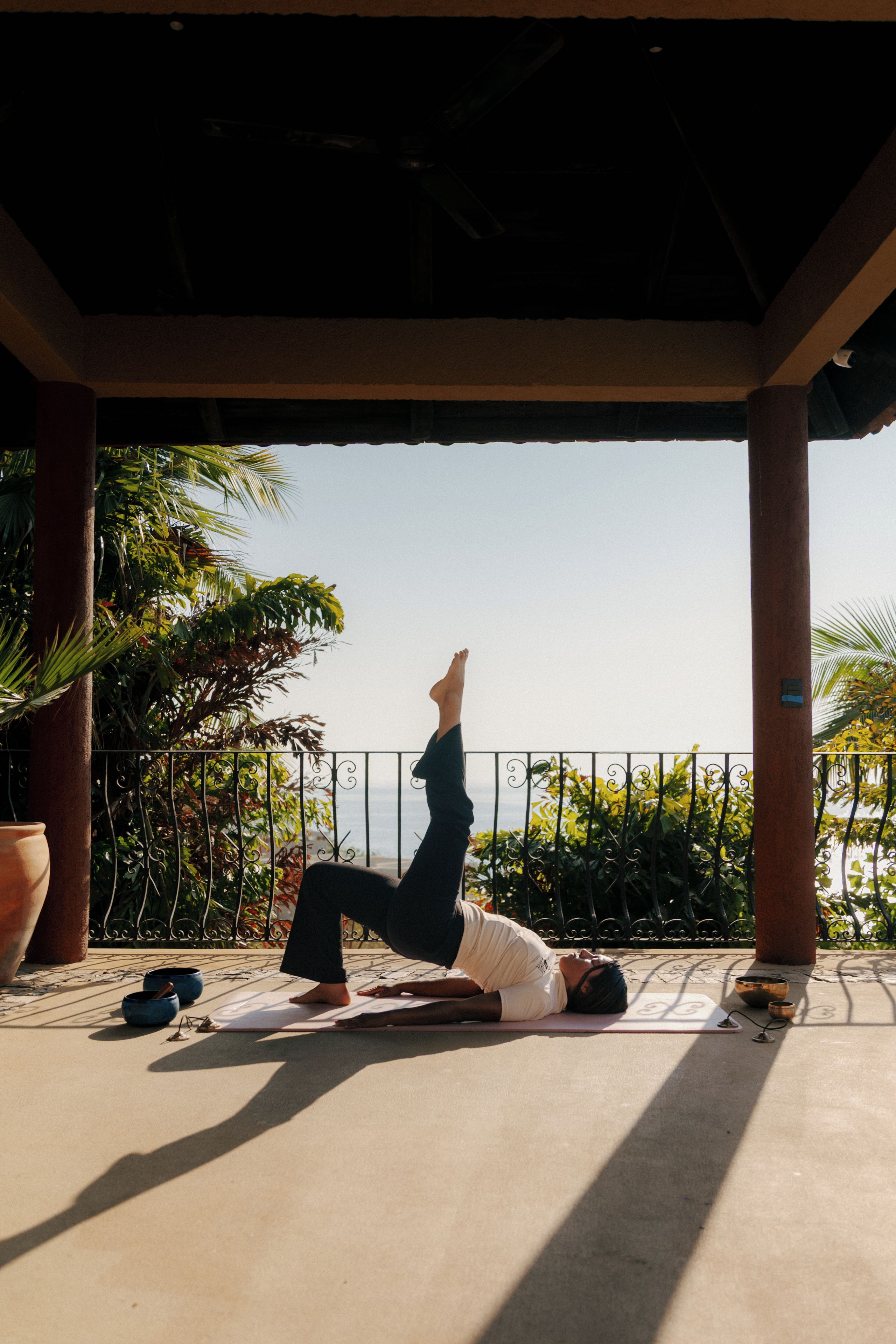 A person performing a yoga pose on a mat outdoors with a railing and plants in the background.