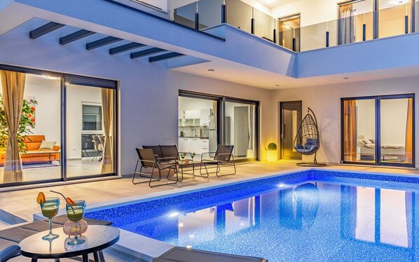 Una piscina interior moderna con cerramiento de vidrio, un comedor con asientos exteriores y una cocina visible a través de amplios ventanales correderas.