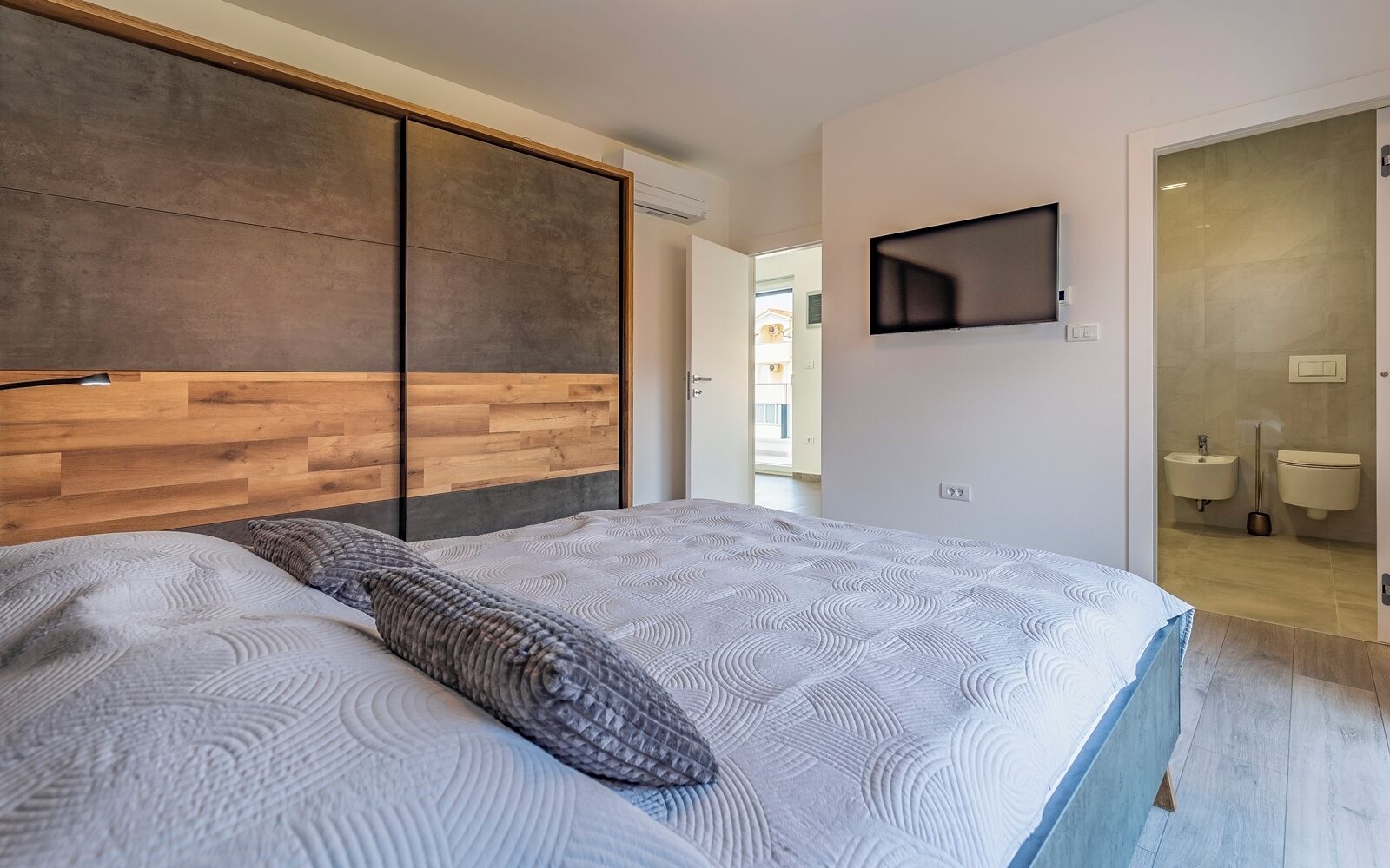 Un dormitorio con cama, cabecera de madera, televisor montado en la pared y baño a la vista a través de una puerta abierta.