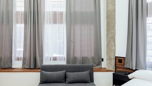 Habitació moderna amb un sofà gris, un llit i una finestra amb cortines fines.