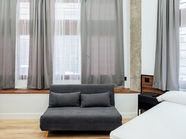 Ein modernes Zimmer mit einem grauen Sofa, einem Bett und einem Fenster mit durchsichtigen Vorhängen.