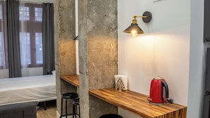 Habitació d'hotel senzilla amb llit, zona de bar de fusta, tetera vermella i lluminària penjada a la paret.