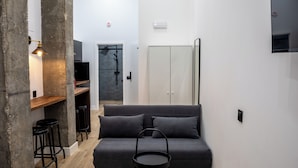 Un espai vital modern i compacte amb un sofà gris, una taula rodona petita, un televisor fixat a la paret i una zona de dutxa.