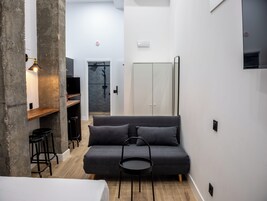 Ein modernes, kompaktes Wohnzimmer mit grauem Sofa, einem kleinen runden Tisch, einem an der Wand montierten Fernseher und einer Duschkabine.