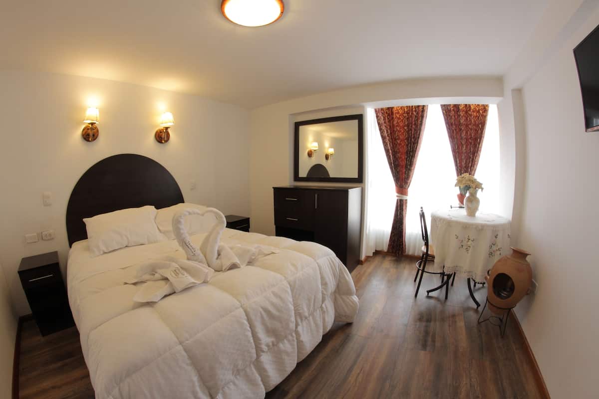 Habitación de hotel con cama, mesitas de noche, cómoda, espejo, una mesa redonda con un florero y cortinas.