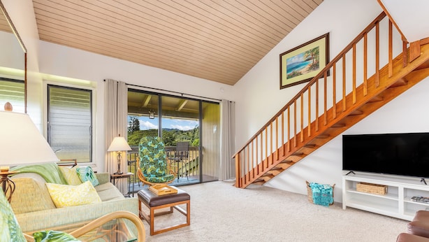 A modern living room with a wooden staircase, a large flat-screen TV, and a view of a balcony with outdoor furniture. - "Da Koa Bar" Paradise at Turtle Bay -Trade Winds, Beaches, Sunshine Rain, Resort (Kahuku)