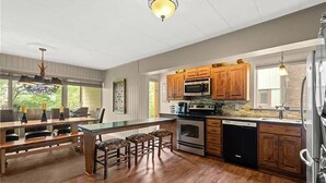 A modern kitchen with wooden cabinets, a central island with bar stools, stainless steel appliances, and a large window providing natural light. - Cozy Condo at Seven Springs
 (Champion)