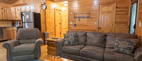 Interior de una cabaña de madera con sala de estar, un sofá, un sillón reclinable, una mesita de centro y una zona de cocina con refrigerador.
