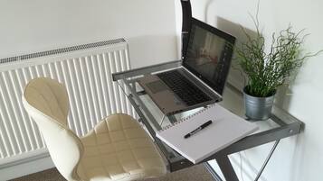 A workspace with a laptop on a glass desk, a cushioned chair, a potted plant, and a radiator in the background.