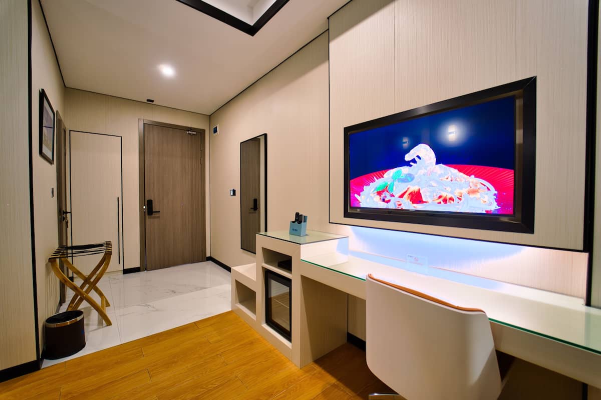 A modern hotel room with a large flat-screen TV mounted on the wall, a glass desk, and a wooden floor.