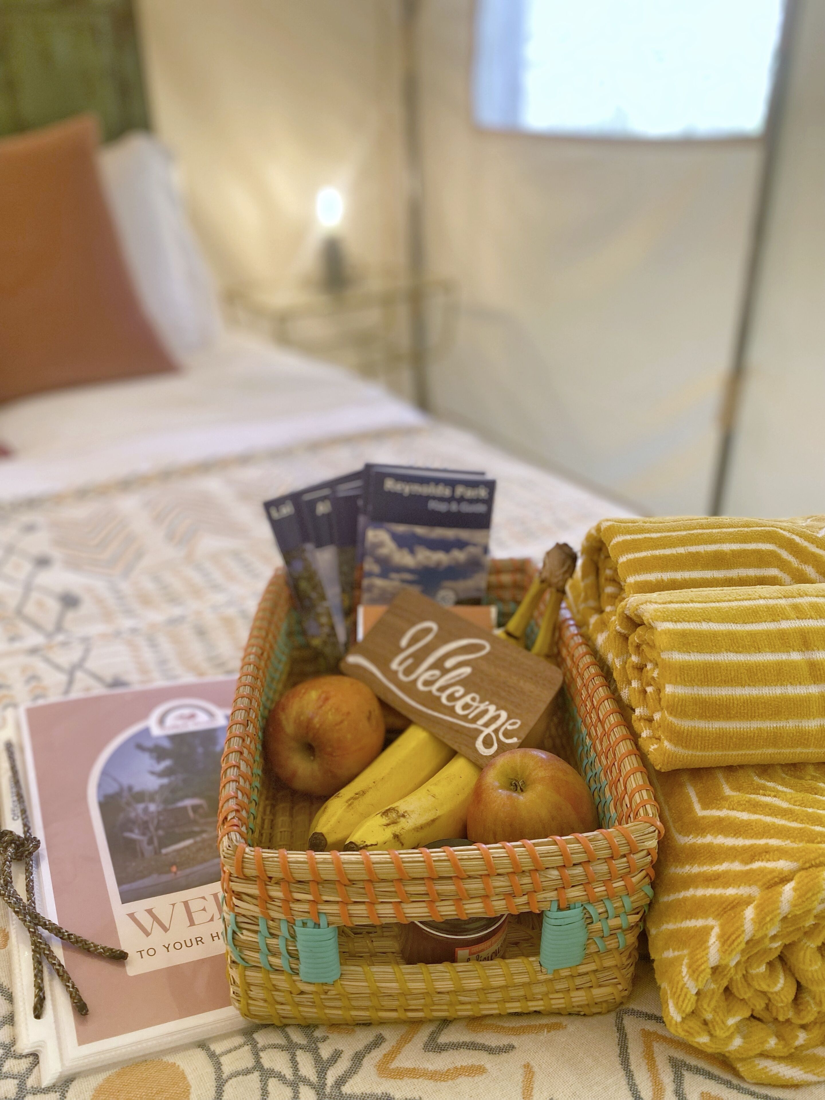 Una cama con una tarjeta de bienvenida, una cesta de mimbre con frutas y una toalla doblada sobre el cubrecolchón.