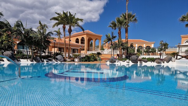 A clear blue swimming pool with tiled edges, surrounded by palm trees and a building with arched windows and a tiled roof. - Breathtaking sea view, only 100 m to the sea and the beach, heated pool, wifi (Playa de la Américas)