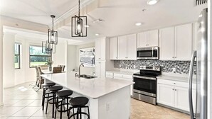 Une cuisine moderne avec des meubles blancs, un grand îlot central doté de tabourets, des appareils en acier inoxydable et un éclairage suspendu.