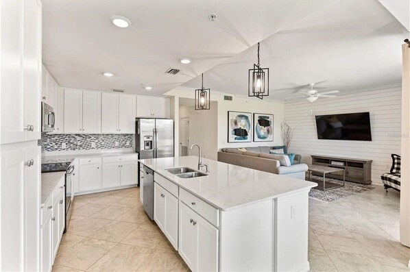 A modern kitchen with white cabinetry, a large island with a sink, and a dining area with a sofa and TV. - Stunning 2BR/2BA Unit overlooking the scenic Lakewood National Golf Course (Lakewood Ranch)