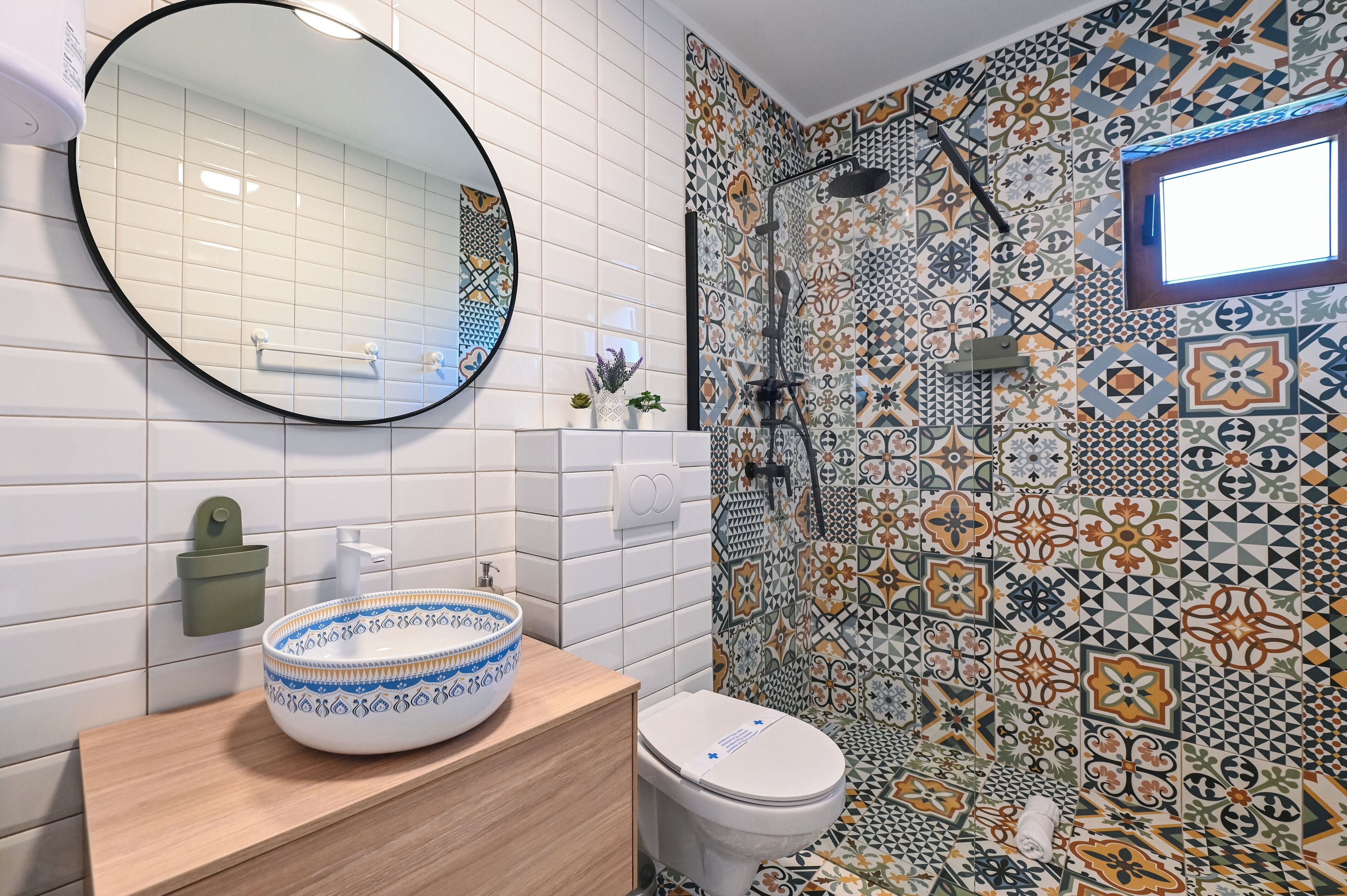 A bathroom with a round mirror, a white toilet, a wooden vanity, and a patterned shower wall.