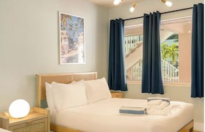 A hotel room with a bed, a nightstand with a lamp, a window with curtains, and a painting on the wall.