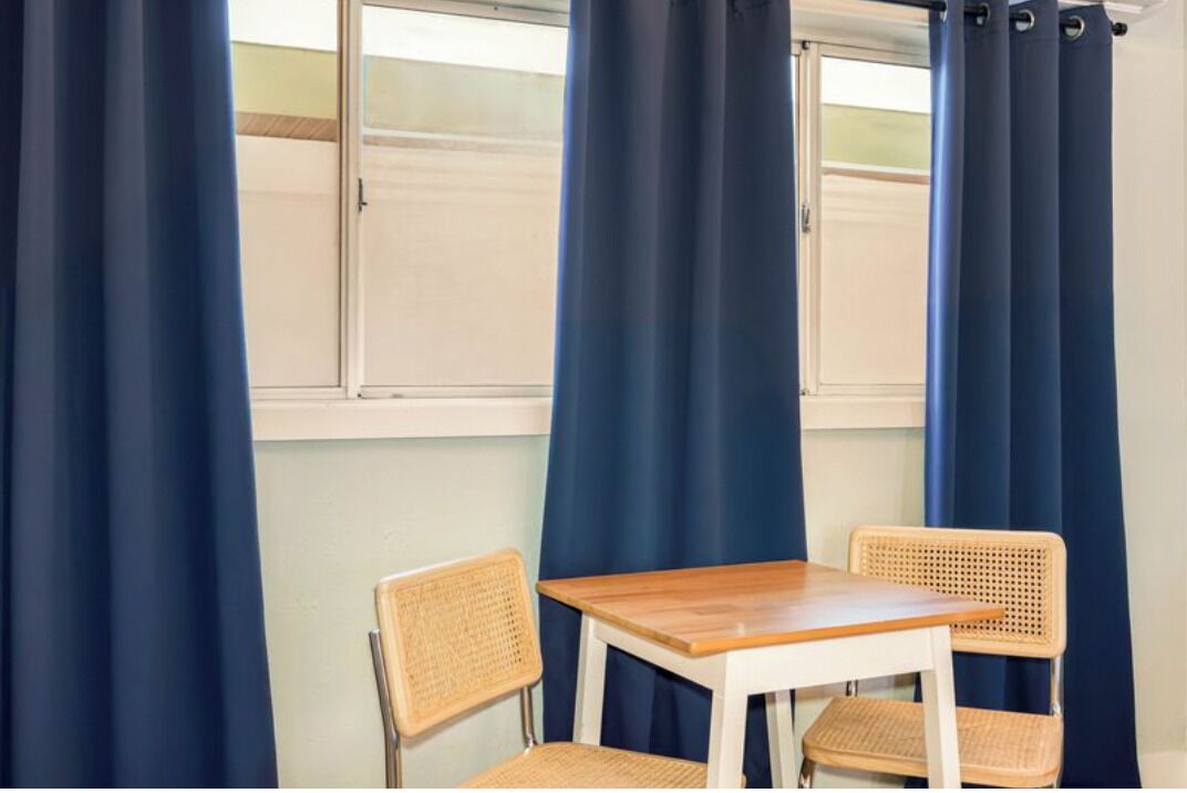 Een kamer met een tafel en twee rieten stoelen, een raam met gordijnen en uitzicht op een gebouw.