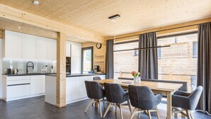 Une cuisine moderne avec des meubles blancs et un îlot central en bois, un coin repas avec une table en bois et quatre chaises, et de grandes fenêtres avec des rideaux.