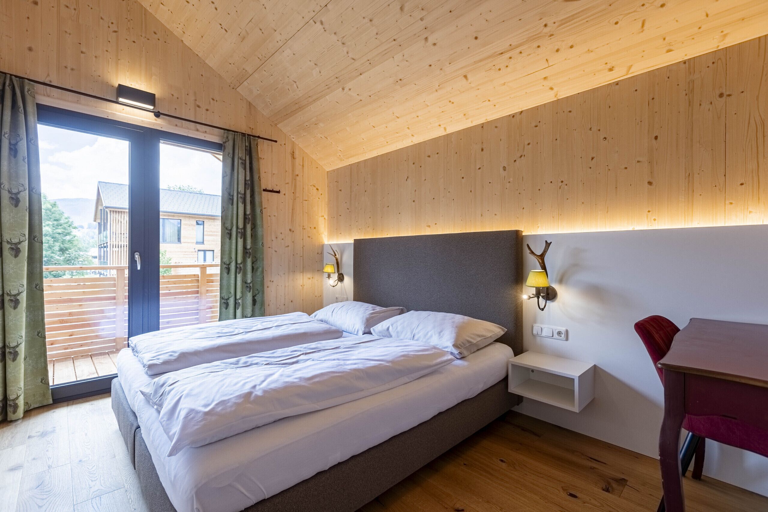 Un dormitorio con techo de madera, una cama con ropa de cama blanca, una mesita de noche con lámpara y vistas a un edificio.