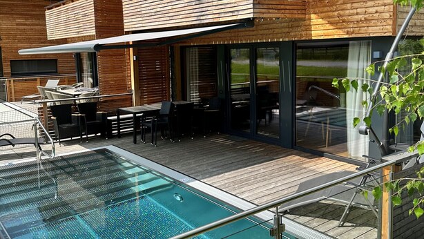 Ein modernes Holzhaus mit einem gläsernen Schwimmteich, Sitzgelegenheiten im Freien und einer überdachten Terrassenfläche.