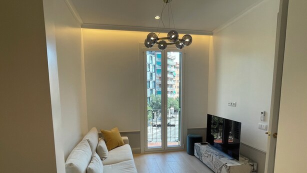 A spacious living room with a white sofa, a TV on a marble stand, and a large window offering a view of the city. - Apartment Luxury one bed one Bathroom (Barcelona)