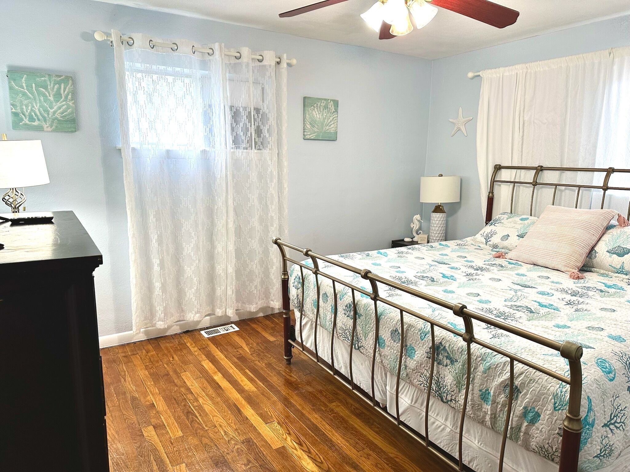 A bedroom with a metal bed frame, a wooden floor, a ceiling fan, and a window with sheer curtains.