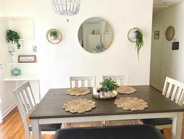 A dining area with a wooden table, white chairs, and woven placemats. There are potted plants on the walls and a chandelier above the table. - Cozy Ranch Home, Centrally Located near Chicks Beach! (Virginia Beach)