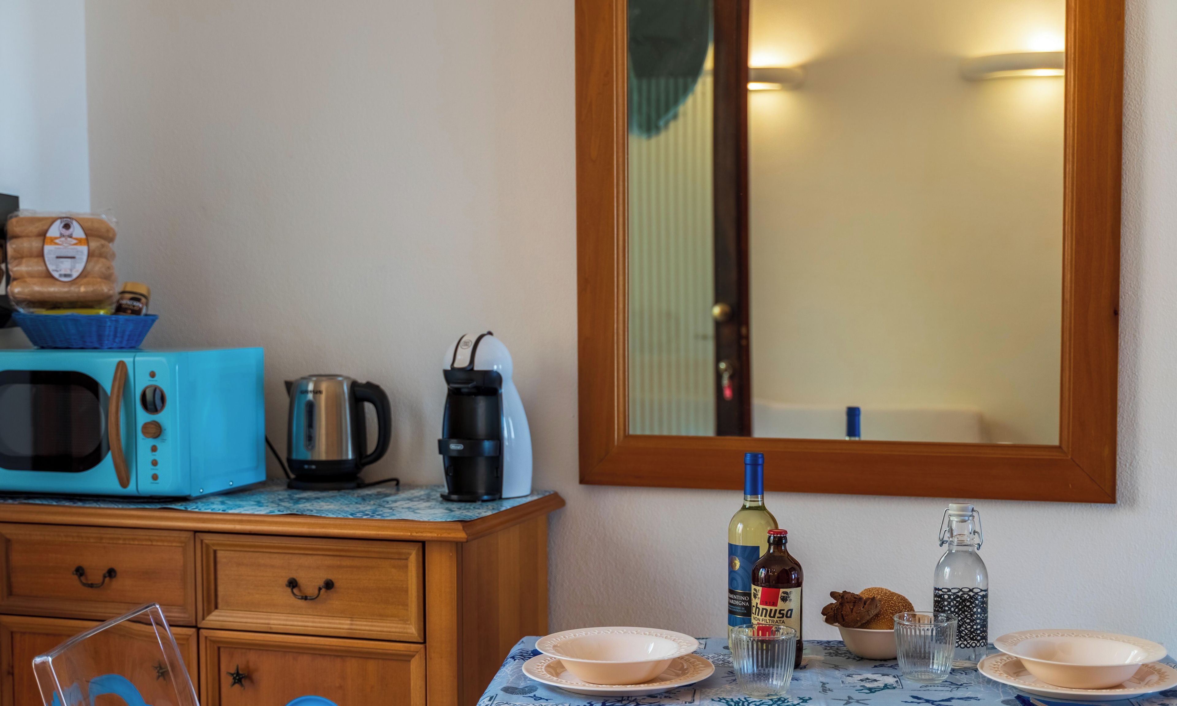 Eine Küchenzeile mit Mikrowelle, Wasserkocher und Kaffeemaschine auf einer Arbeitsfläche. Ein Tisch mit Tellern, einer Flasche und einem Glas ist ebenfalls vorhanden.