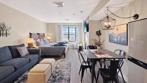 Quarto de hotel moderno com sofá, cama, mesa de jantar, cadeiras e uma janela grande.