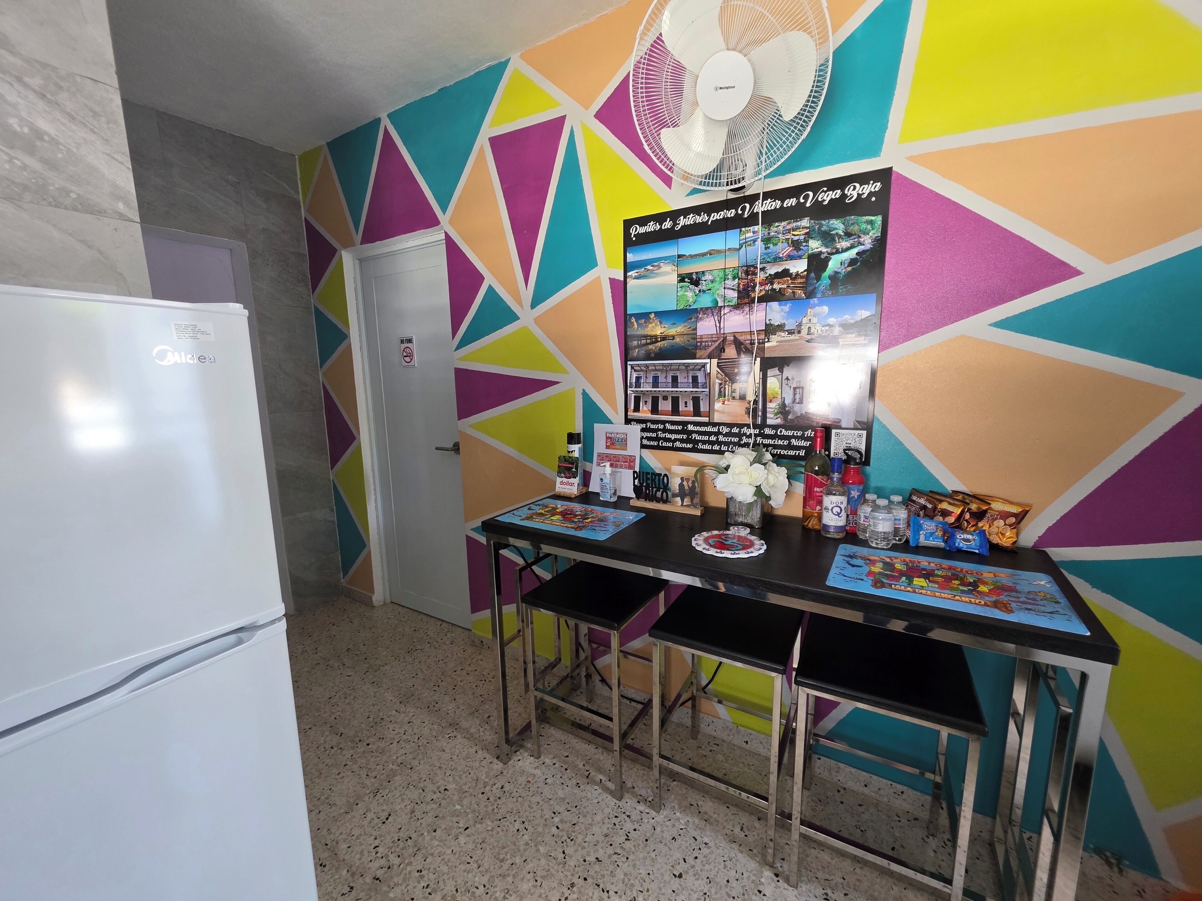 Una cocina con refrigerador, una mesa de comedor con sillas y una pared colorida con un cartel.