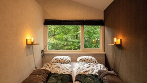 A room with two beds, a window with a view of greenery, and wall-mounted lights.