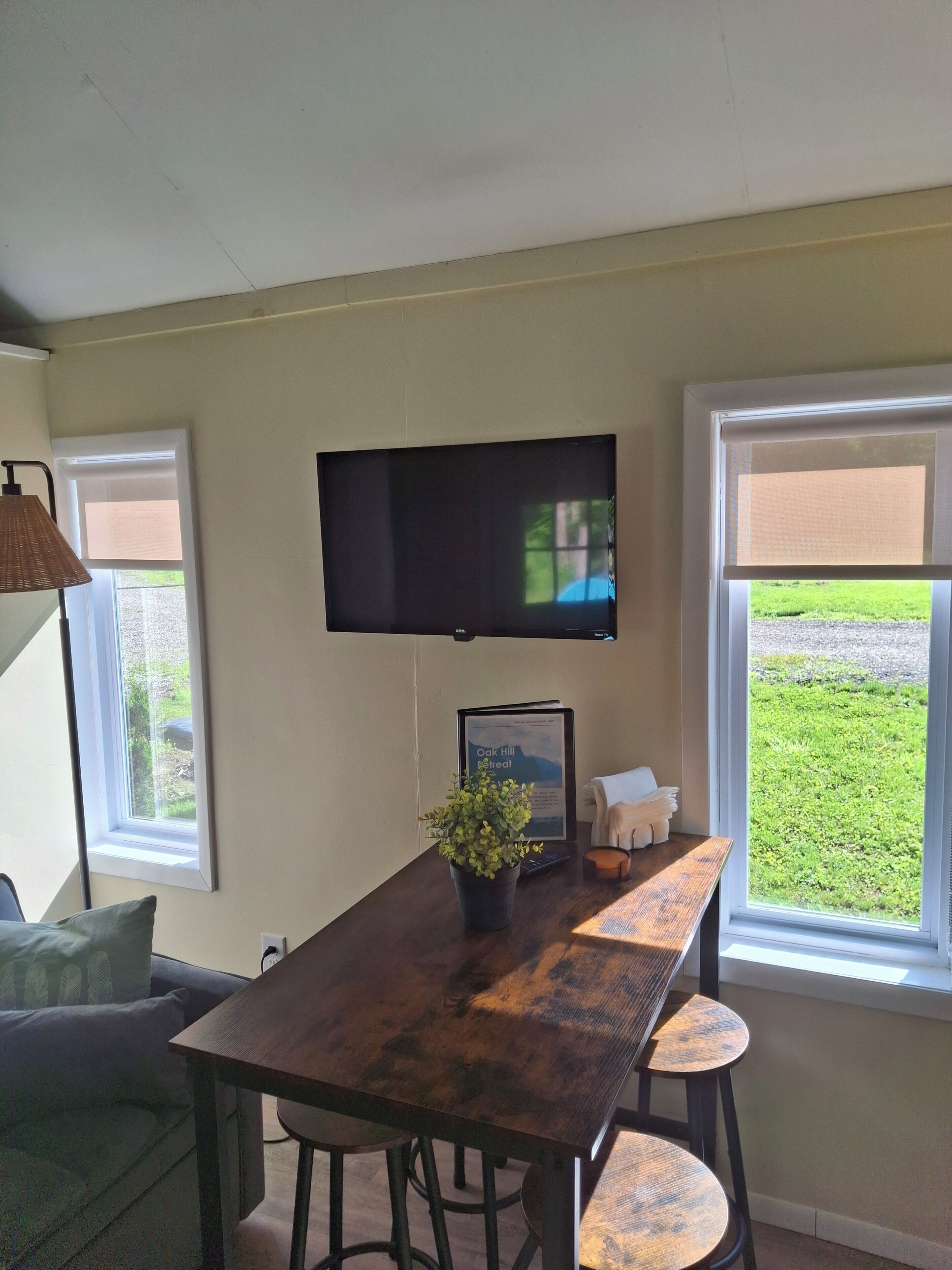 A dining area with a table, chairs, a television mounted above, and windows with a view of greenery.