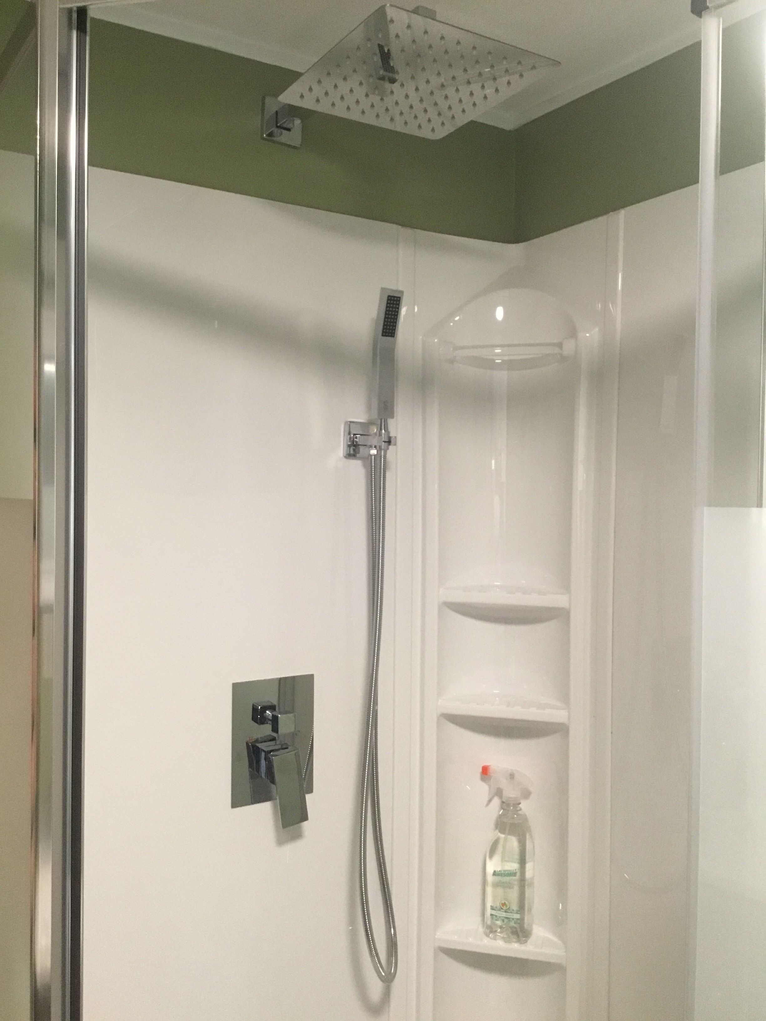 A shower enclosure with a handheld showerhead, a wall-mounted control panel, and a shelf with a bottle of liquid soap.