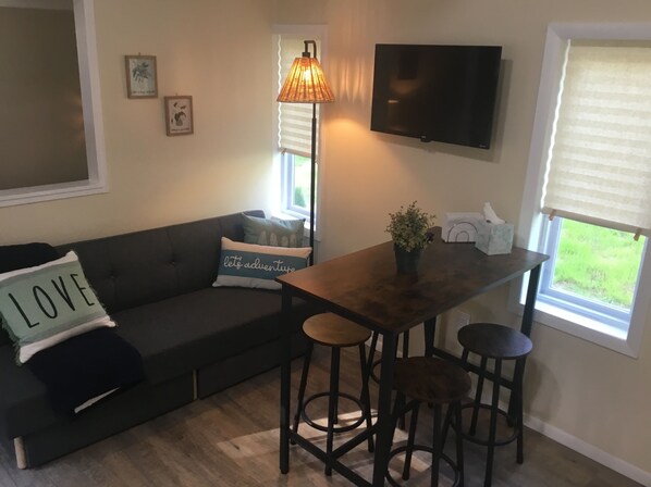 A living room with a dark gray sofa, a wooden coffee table, and two bar stools. There is a television mounted on the wall, a window with blinds, and a potted plant on the table. - Mid-Century Tiny House in the heart of New River Gorge National Park (Oak Hill)