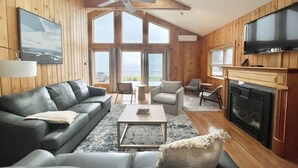 Un salón amplio con un sofá grande, una mesa de centro y una chimenea. Varias ventanas ofrecen luz natural y vistas al exterior.